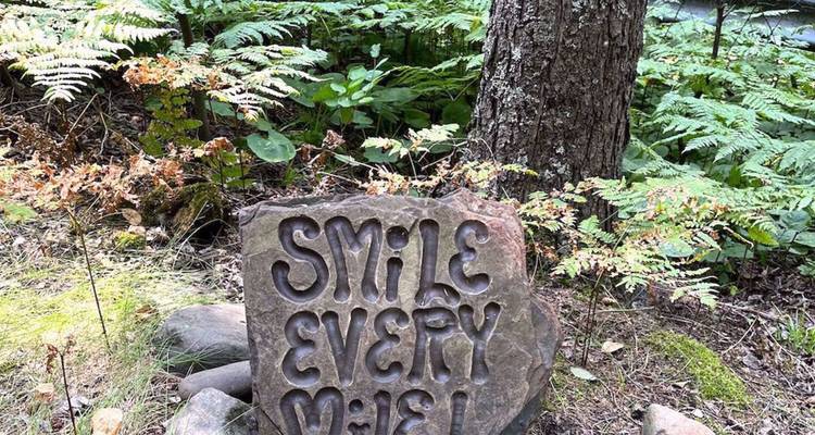 Pierre décorative dans un jardin boisé gravée des mots « Souriez à Chaque Kilomètre ! »