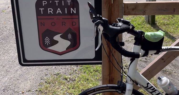 Un vélo s'appuie contre un panneau de sentier portant l'inscription « P'tit Train du Nord » avec un logo de sentier stylisé.