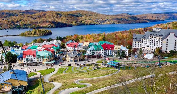 Vue panoramique sur le village coloré de Mont-Tremblant, un grand lac et des collines forestières ondulantes sous un ciel partiellement nuageux.