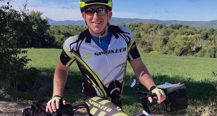 Un cycliste souriante en tenue complète enfourche un vélo sur une route de campagne avec des collines vertes ondulantes en arrière-plan.