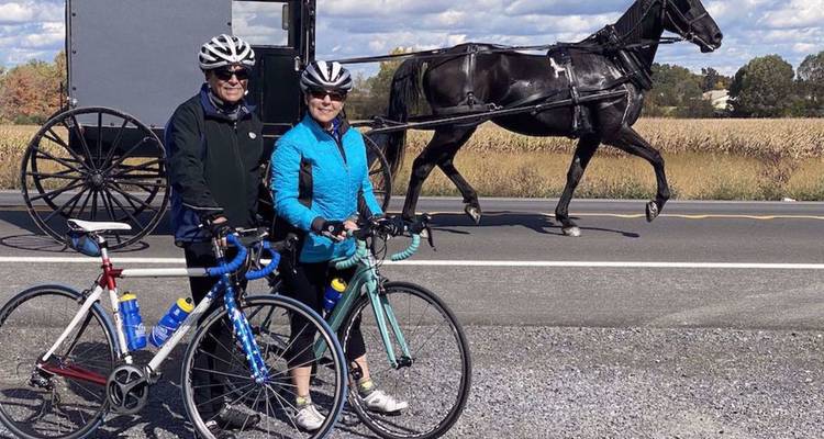 Deux cyclistes posent avec leurs vélos sur le bord d'une route rurale tandis qu'un buggy tiré par un cheval noir passe derrière eux.