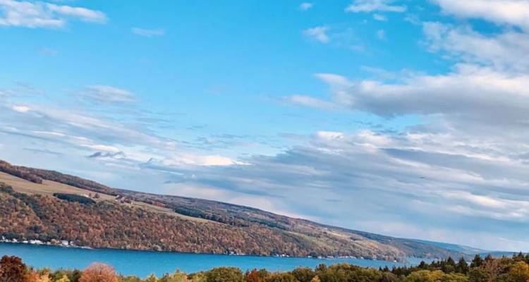 Vue panoramique d'un lac des Finger Lakes bordé de collines vallonnées d'automne sous un ciel lumineux.