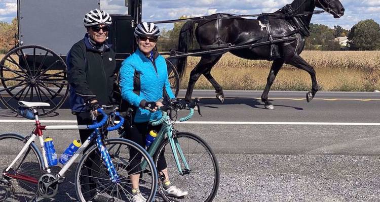 Un couple à vélo pose au bord de la route tandis qu'un cheval et un buggy amish passent derrière.