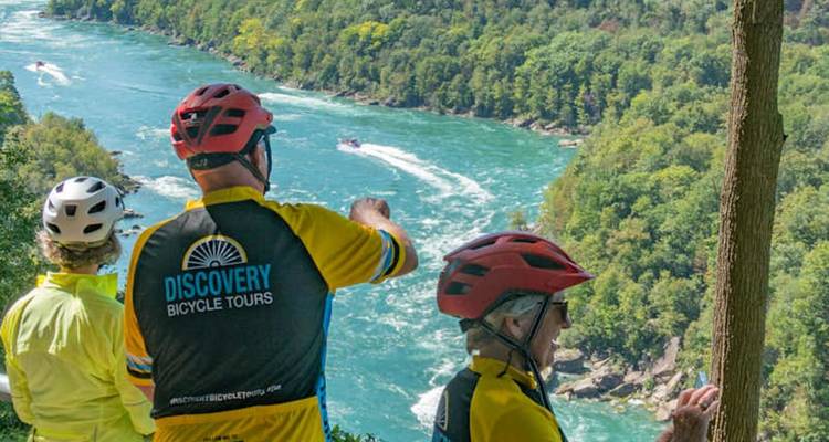 Les cyclistes portant des casques regardent vers le bas une gorge fluviale d'un turquoise profond avec des bateaux à moteur rapides en dessous.