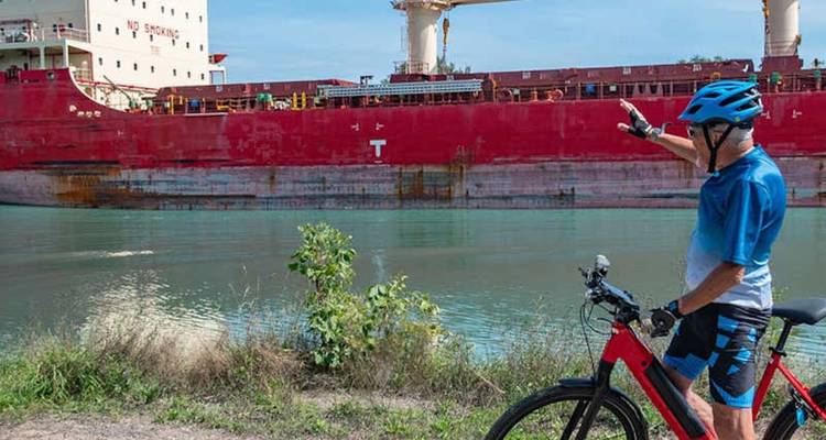 Un cycliste fait un geste en direction d'un énorme navire de cargo rouge qui traverse un canal.