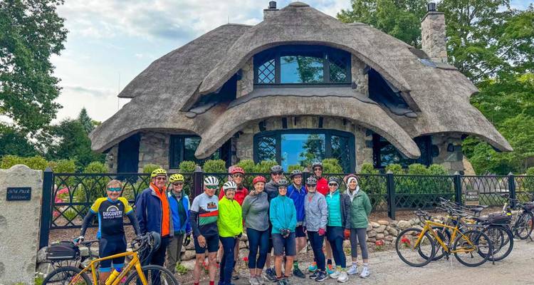 Een grote groep fietsers poseert voor een speels stenen huisje met een dramatisch rieten dak.