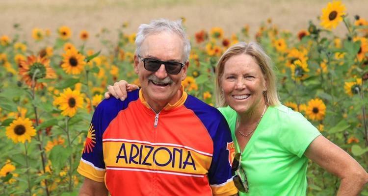 Glimlachend stel staat midden in een veld met hoge zonnebloemen, de man draagt een Arizona wielrenshirt.