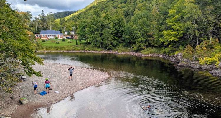 Un petit groupe profite d'une rive de galets tandis qu'une personne nage dans les eaux calmes à côté d'une vallée boisée.