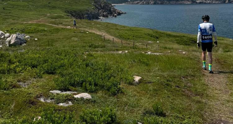 Un participant portant un dossard d'événement marche sur un sentier côtier herbeux surplombant une eau d'un bleu profond.