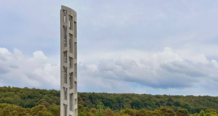 Een hoog, modern betonnen gedenktoren steekt uit boven een groen woud onder een bewolkte hemel.