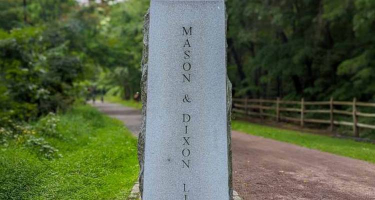 Een granietmarker met "Mason & Dixon Line" gegraveerd staat naast een met bomen omzoomd grindpad.