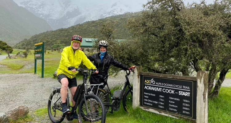 Deux cyclistes vêtus de vestes imperméables posent avec leurs vélos devant le panneau officiel du départ de la piste cyclable Alps 2 Ocean, au milieu de collines brumeuses.