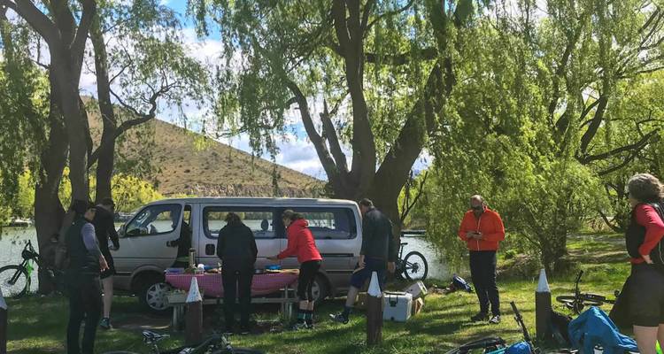Un groupe de cyclistes se réunit pour un pique-nique près d'une camionnette sous des arbres ombragés en bord de rivière.