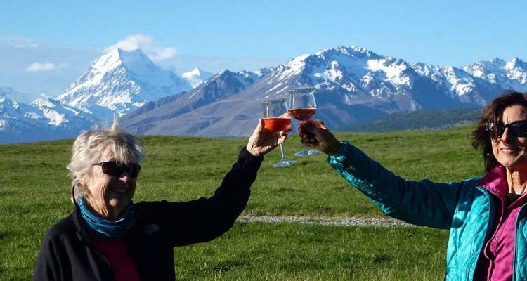 Deux femmes trinquent avec du vin rosé sur fond de pics enneigés et de pâturages ouverts.