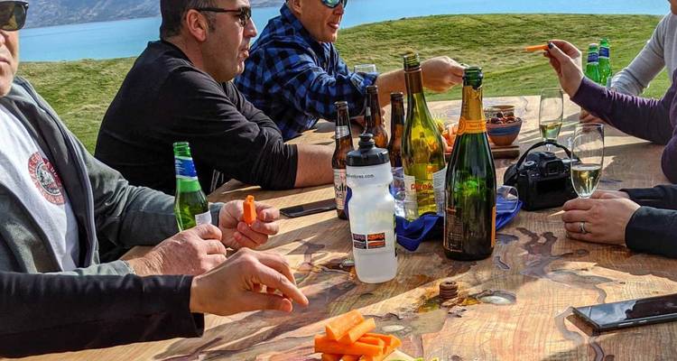 Un groupe déguste des boissons et des collations autour d'une table en bois rustique donnant sur un lac turquoise.