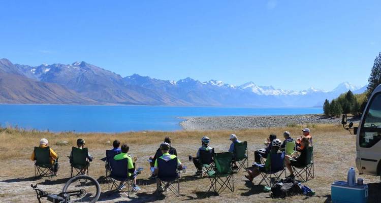 Grand groupe assis sur des chaises de camping près d'un lac alpin d'un bleu éclatant face à un panorama de montagnes enneigées.