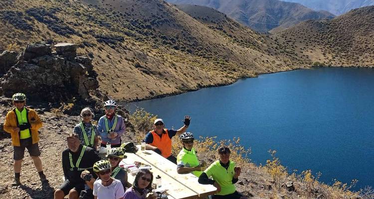 Un groupe de cyclistes faisant la fête salue depuis une table de pique-nique au sommet d'une falaise surplombant un lac d'un bleu éclatant dans des collines escarpées.