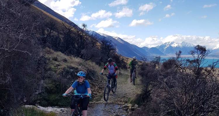 Les vététistes naviguent sur un sentier accidenté dominant un lac alpin turquoise encadré par des arbustes sans feuilles.