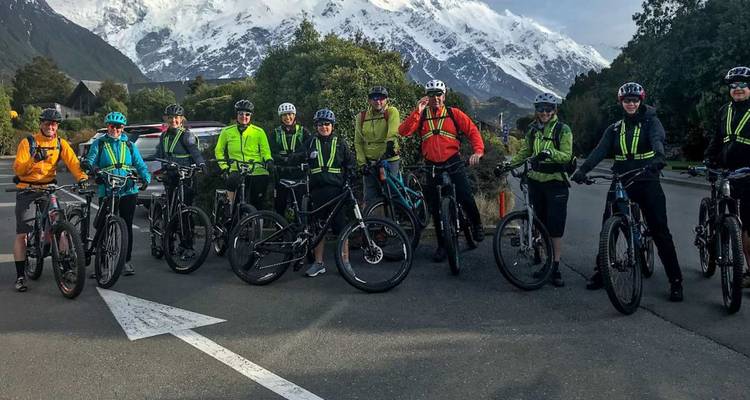 Un grand groupe de cyclistes pose avec leurs vélos sur une rue de village sous des pics enneigés imposants.