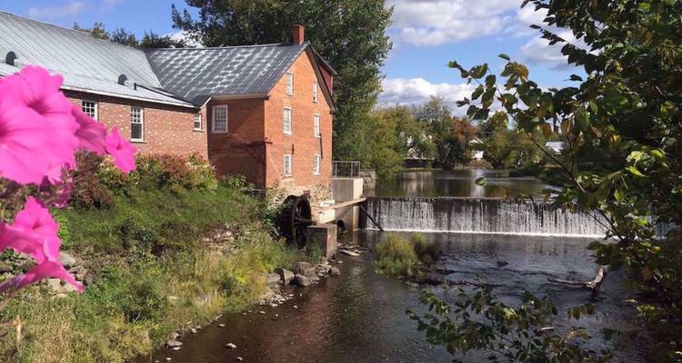 Moulin historique en brique situé à côté d'une petite cascade sur une rivière entourée de fleurs et d'arbres