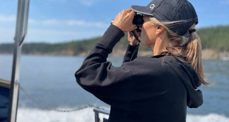 Une femme à bord d'un bateau scrute l'horizon à l'aide de jumelles tandis que le sillage tourbillonne derrière elle.