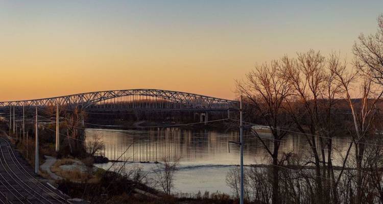 Spoorwegbrug die een reflectieve rivier overspant bij schemering met kale bomen afgetekend tegen een oranje lucht