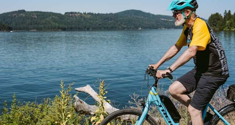 Un cycliste âgé chevauche un vélo électrique le long de la rive d'un grand lac bleu aux collines boisées.