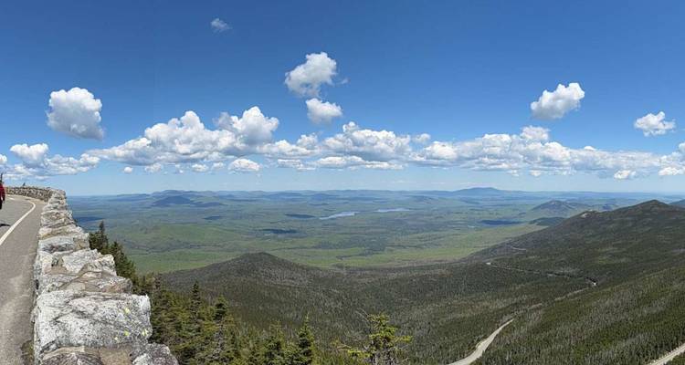Point de vue panoramique en bordure de route surplombant des montagnes vertes ondulantes et des lacs lointains sous des nuages épars.