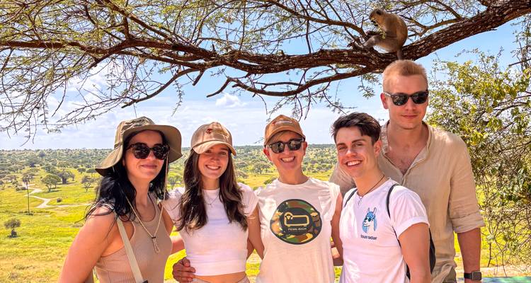 Grupo sonriente de jóvenes viajeros posando bajo un árbol mientras un mono está sentado encima de ellos.