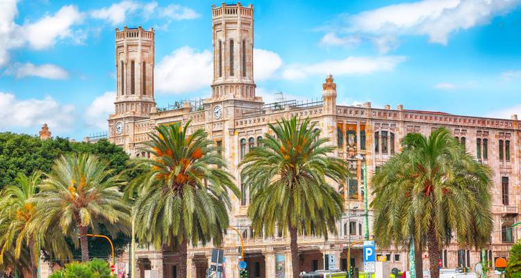 Edificio municipal de arenisca rosa histórica con dos torres de reloj gemelas enmarcadas por altas palmeras bajo un cielo brillante en Cagliari.