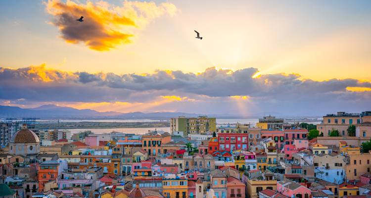 Los rayos del atardecer se abren paso entre las nubes sobre los techos multicolores de Cagliari con pájaros volando en lo alto.