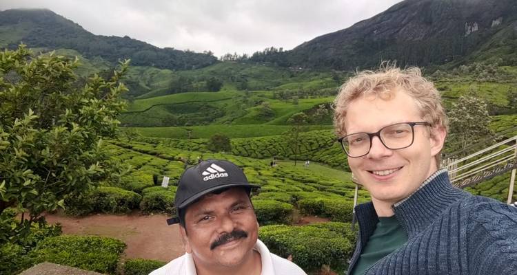 Selfie von zwei Männern, die lächeln, mit ausgedehnten terrassierten Teefeldern und Hügeln im Hintergrund.