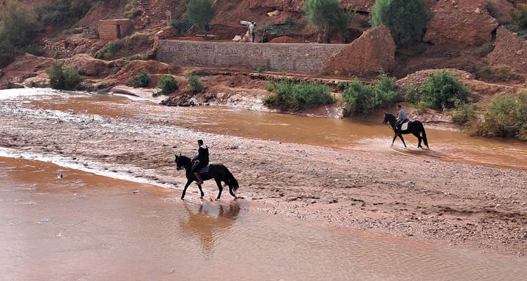 Ruiters te paard steken een ondiep modderig riviert je over dat omgeven wordt door rode aarden terrassen en schaarse begroeiing.