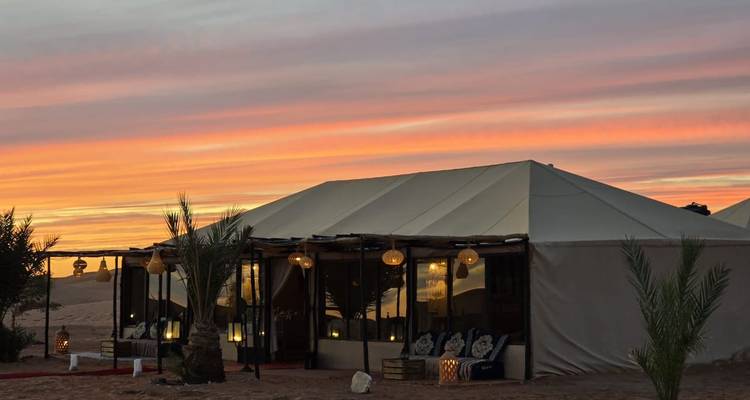 Luxe woestijntentenkamp verlicht bij zonsondergang onder een gestreepte oranje en roze hemel.