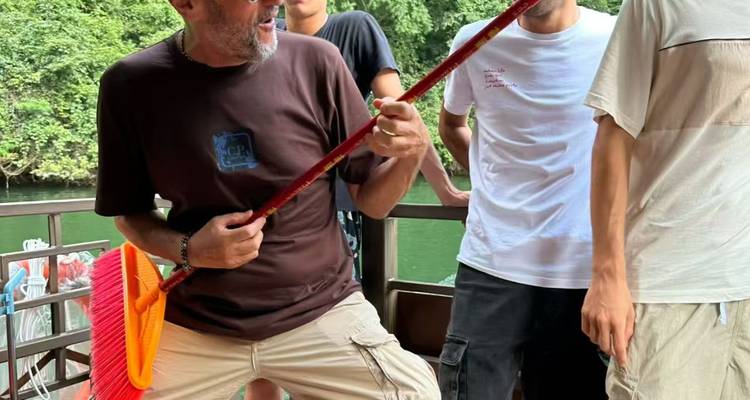 Un hombre toca de forma juguetona una escoba como si fuera una guitarra en la cubierta de un bote mientras sus compañeros ríen junto a un telón de fondo de río verde.