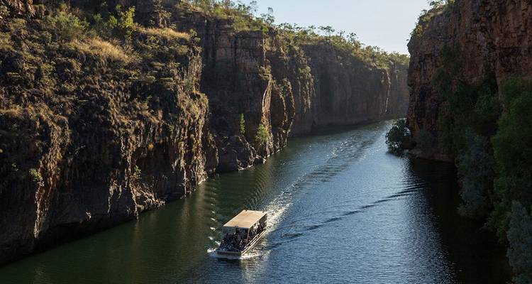 Un bateau de tourisme transportant des passagers glisse à travers une gorge étroite aux falaises rouges escarpées et aux eaux fluviales calmes sous la lumière de fin d'après-midi.
