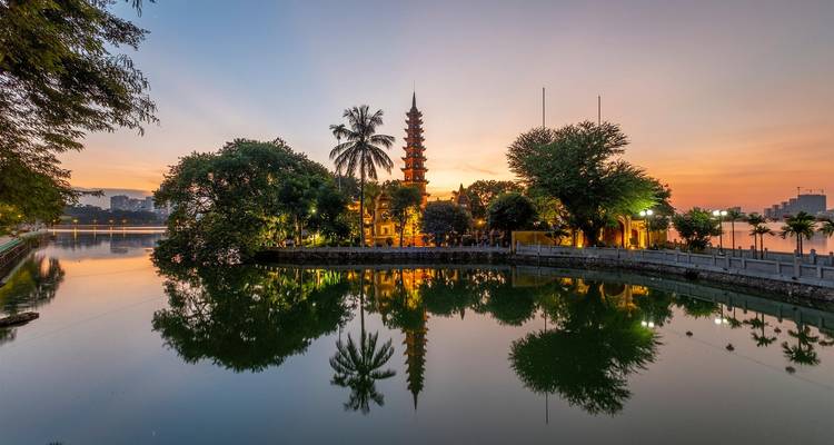 Pagode Tran Quoc et reflet au coucher du soleil sur le Lac de l'Ouest avec un ciel orangé et des arbres luxuriants.
