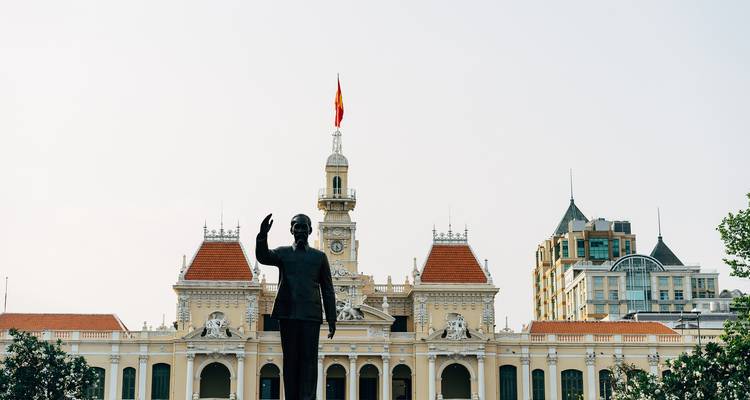 Statue de Hô Chi Minh saluant dans une place officielle devant l'Hôtel de Ville de l'époque coloniale surmonté d'un drapeau national.