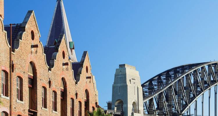 Historische Backsteinmagazine von The Rocks neben den Stahlbögen der Sydney Harbour Bridge unter klarem Himmel.