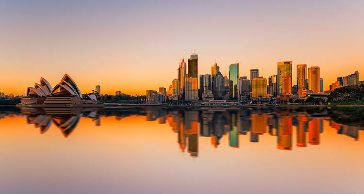 Goldener Sonnenaufgang über der Skyline von Sydney mit Opernhaus und Spiegelungen auf ruhigen Hafengewässern