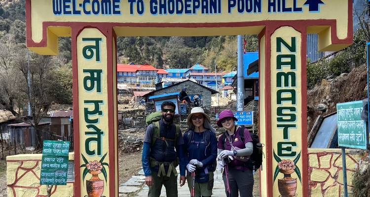 Trois trekkeurs souriants sous l'arche colorée « Bienvenue à Ghodapani Poon Hill » dans l'Himalaya.