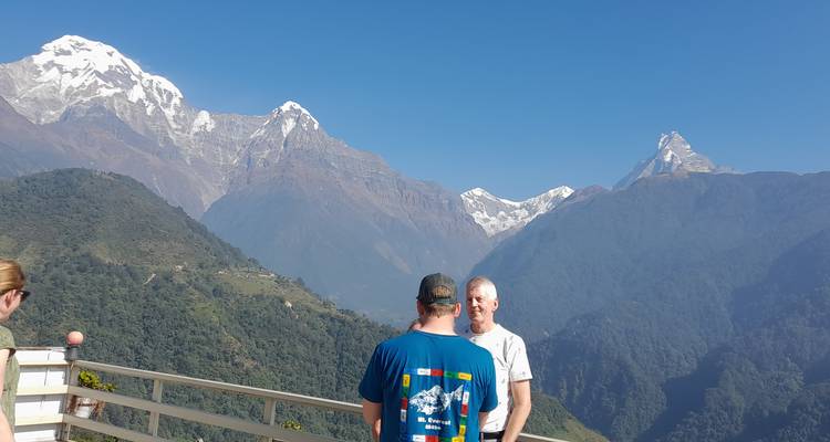 Deux voyageurs discutant sur un balcon avec vue panoramique sur les pics de l'Annapurna sous un ciel bleu dégagé.