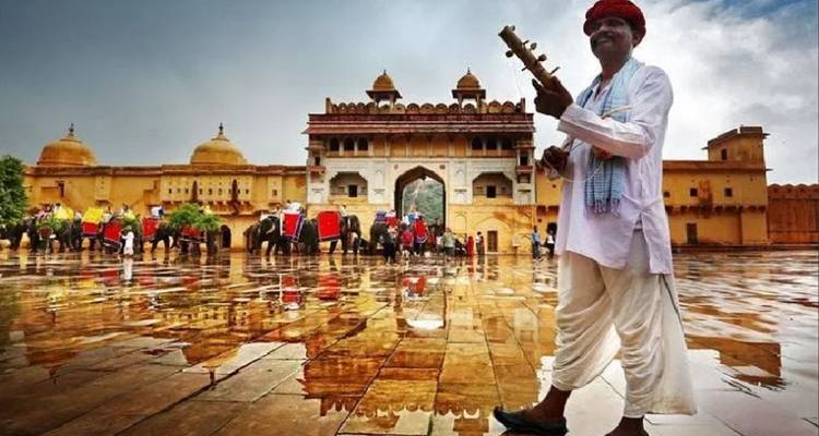 Rajasthani-Musiker in traditioneller Kleidung, der über den regennassen Innenhof des Amer Fort geht, mit Elefanten im Hintergrund.
