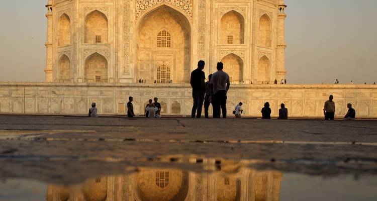 Voyageurs assis et debout près d'une flaque d'eau réfléchissante avec le Taj Mahal illuminé par le soleil en arrière-plan.