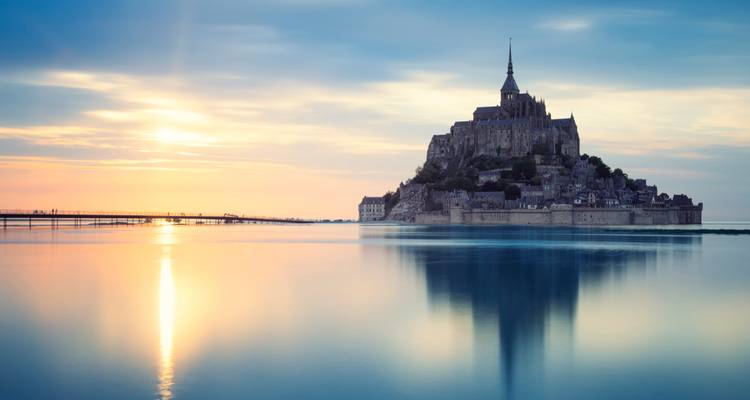 Mont Saint-Michel reflété dans une eau tranquille lors d'un lever de soleil aux teintes pastel délicates.