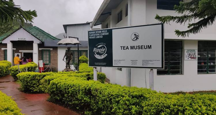Eingang des Ripple Tea Museum in Munnar mit Besuchern, die in der Nähe von Beschilderung und gepflegten Hecken stehen.