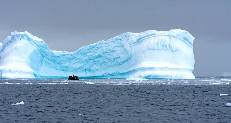 Ein massiver blauer Eisberg überragt ein kleines Zodiac-Boot auf dem grauen Südlichen Ozean.
