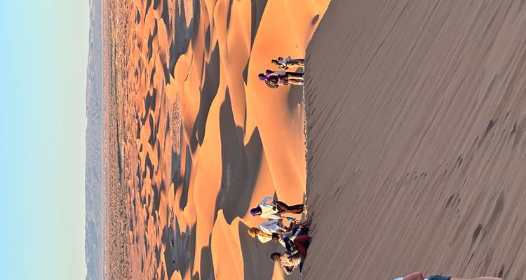 Des voyageurs escaladant une haute dune orange dans le vaste désert de l'Erg Chigaga en fin d'après-midi.