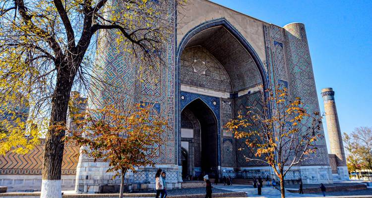 Portal de azulejos majestuoso y minarete cilíndrico del complejo histórico de Samarcanda con transeúntes.