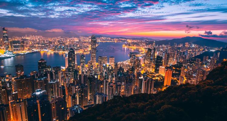 Vista panorámica de los rascacielos de Hong Kong y el puerto iluminados bajo un cielo de atardecer con colores vibrantes, tal como se ve desde el Pico Victoria.